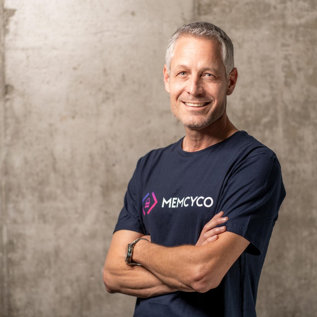 A man with gray hair stands with arms crossed, smiling, wearing a dark blue "MEMCYCO" t-shirt against a plain concrete wall, ready to represent at Cybertech Tel Aviv 2025.